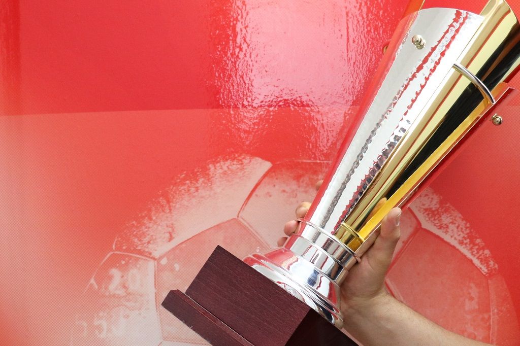 Ein glänzender Silbertrophäe wird von einer Hand gehalten, vor einem roten Hintergrund mit einem Fußballmotiv.