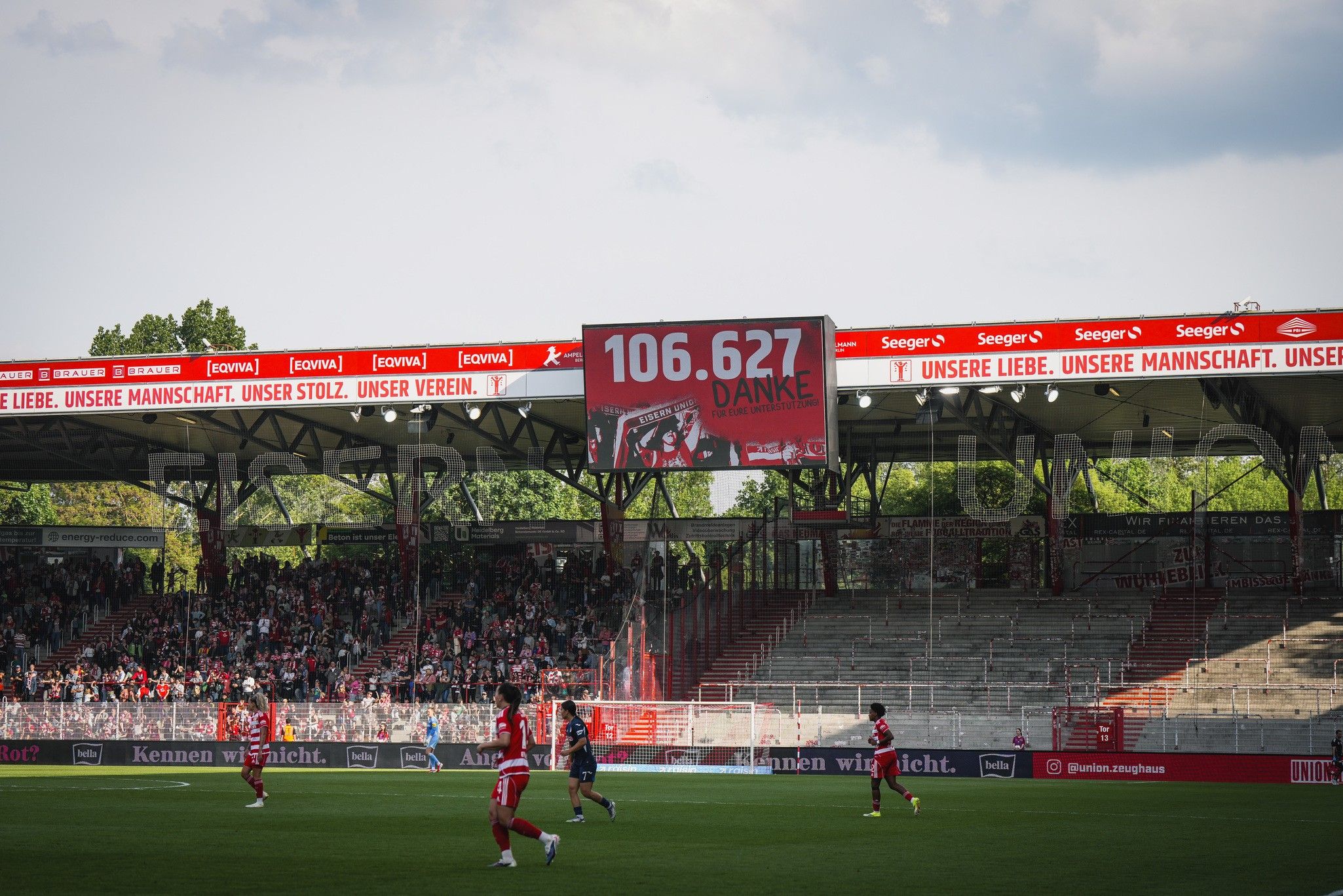 Ein Stadion mit Spielern auf dem Feld, Zuschauern auf den Tribünen und einem großen Bildschirm, der "106.627 DANKEN" anzeigt.