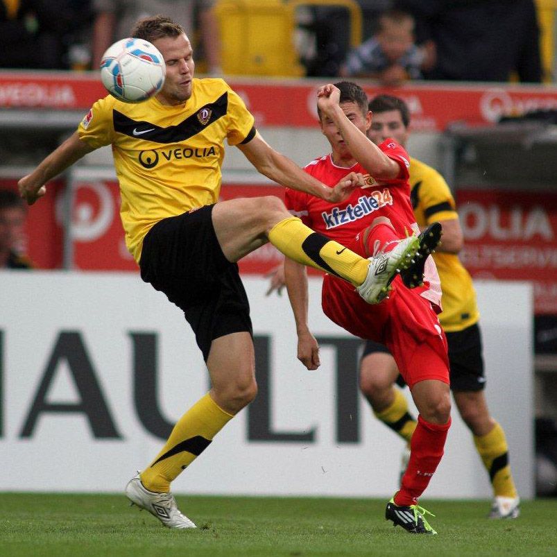 Ein Fußballspieler in gelben und schwarzen Trikots hebt das Bein, während ein anderer Spieler in rot versucht zu verteidigen.