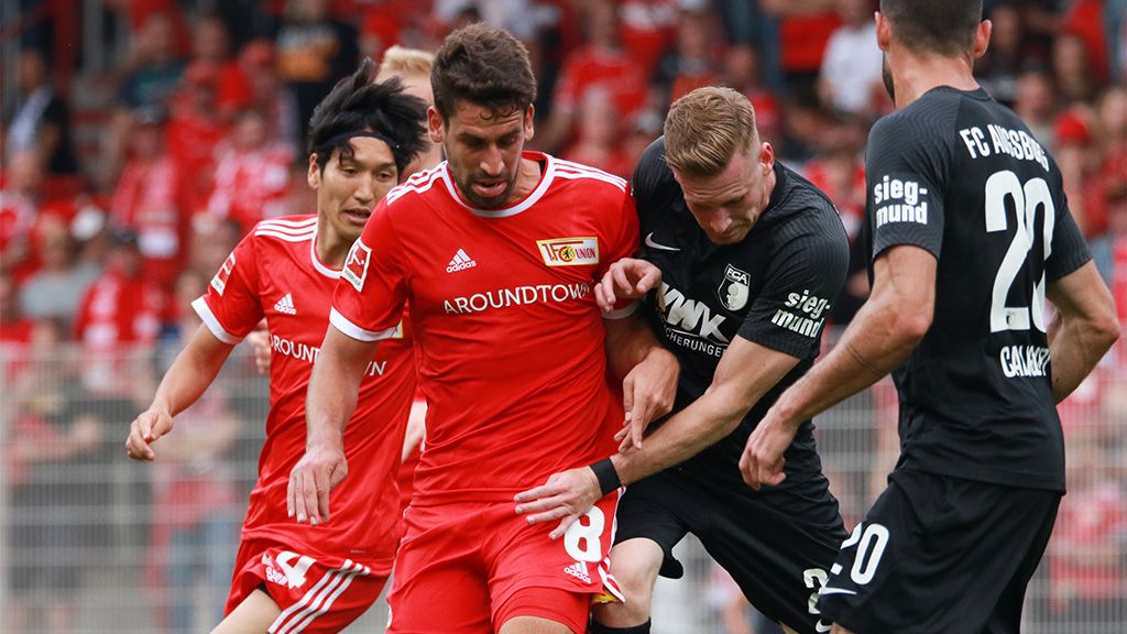 Fußballspiel zwischen zwei Teams, Spieler in roten und schwarzen Trikots kämpfen um den Ball auf dem Spielfeld.