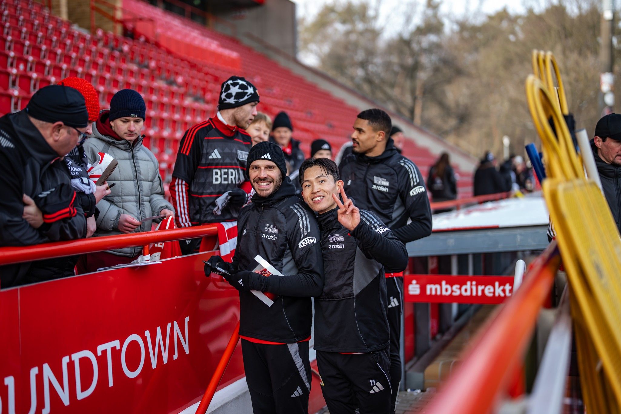 Zwei Fußballspieler in Trainingsanzügen lächeln und posieren mit Fans in einem Stadion. Rote Sitzreihen im Hintergrund.