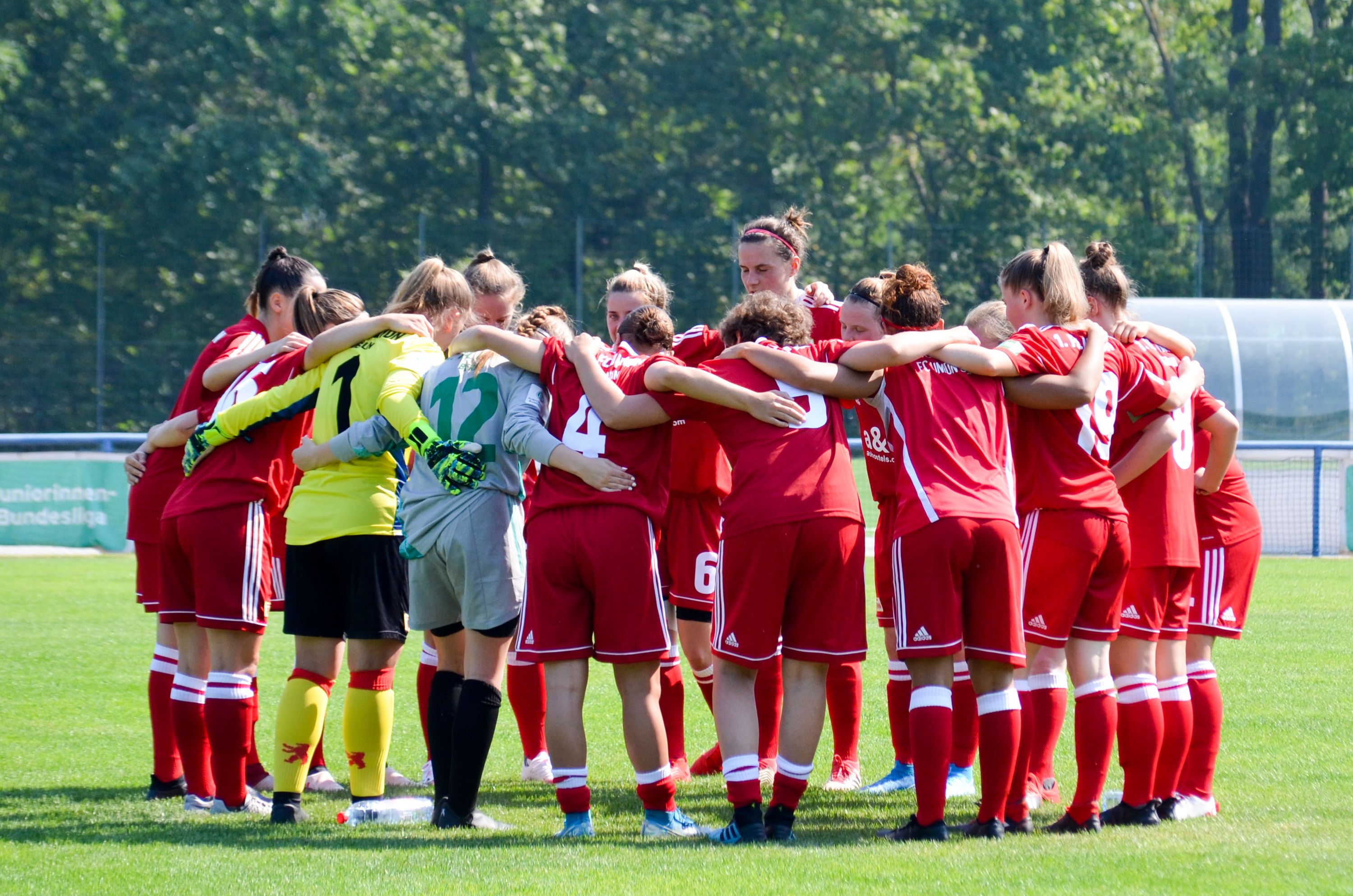 Frauenfußballmannschaft in roten Trikots steht im Kreis und bespricht sich vor einem Spiel auf einem grünen Sportplatz.