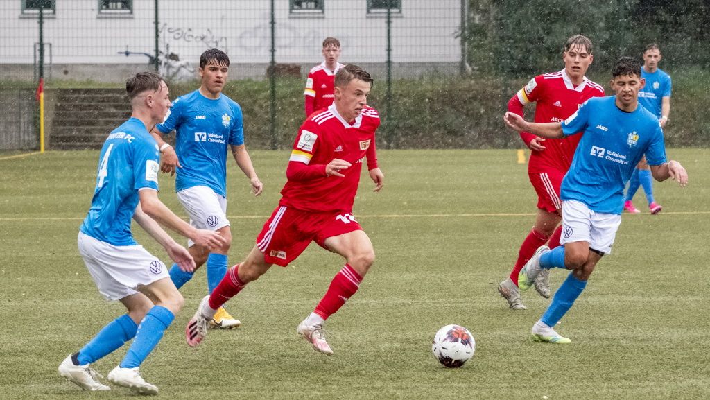 Eine Fußballszene im Regen, mit Spielern in roten und blauen Trikots, die um den Ball kämpfen.