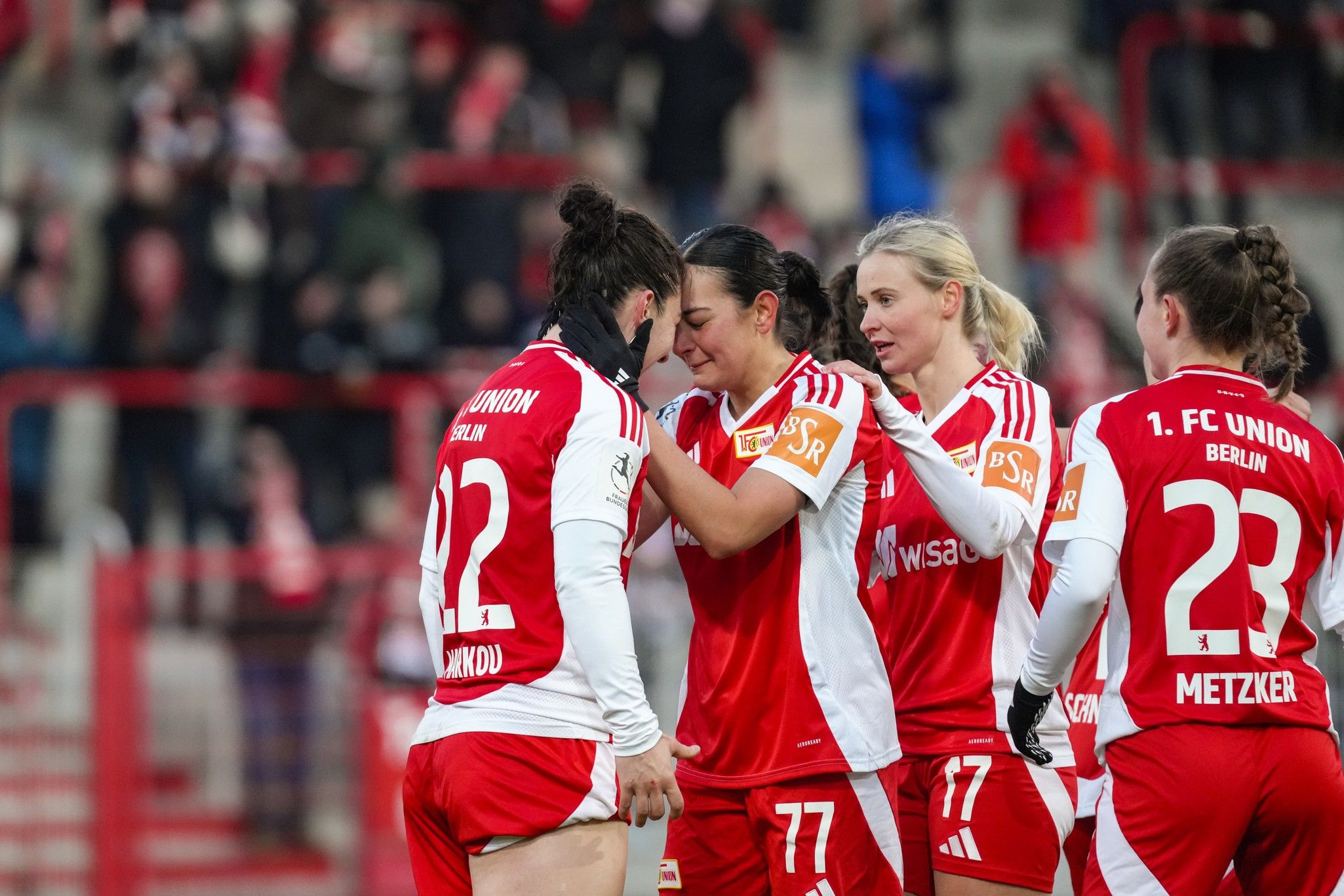 Vier Frauen in roten Fußballtrikots umarmen sich freudig auf einem Spielfeld, während im Hintergrund Fans jubeln.