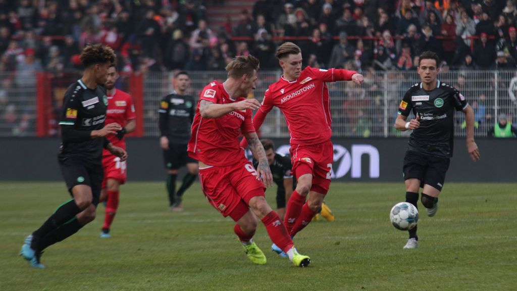 Zwei Spieler in roten Trikots kämpfen um den Ball gegen einen Spieler in grünem Trikot auf einem Fußballfeld. Zuschauer im Hintergrund.