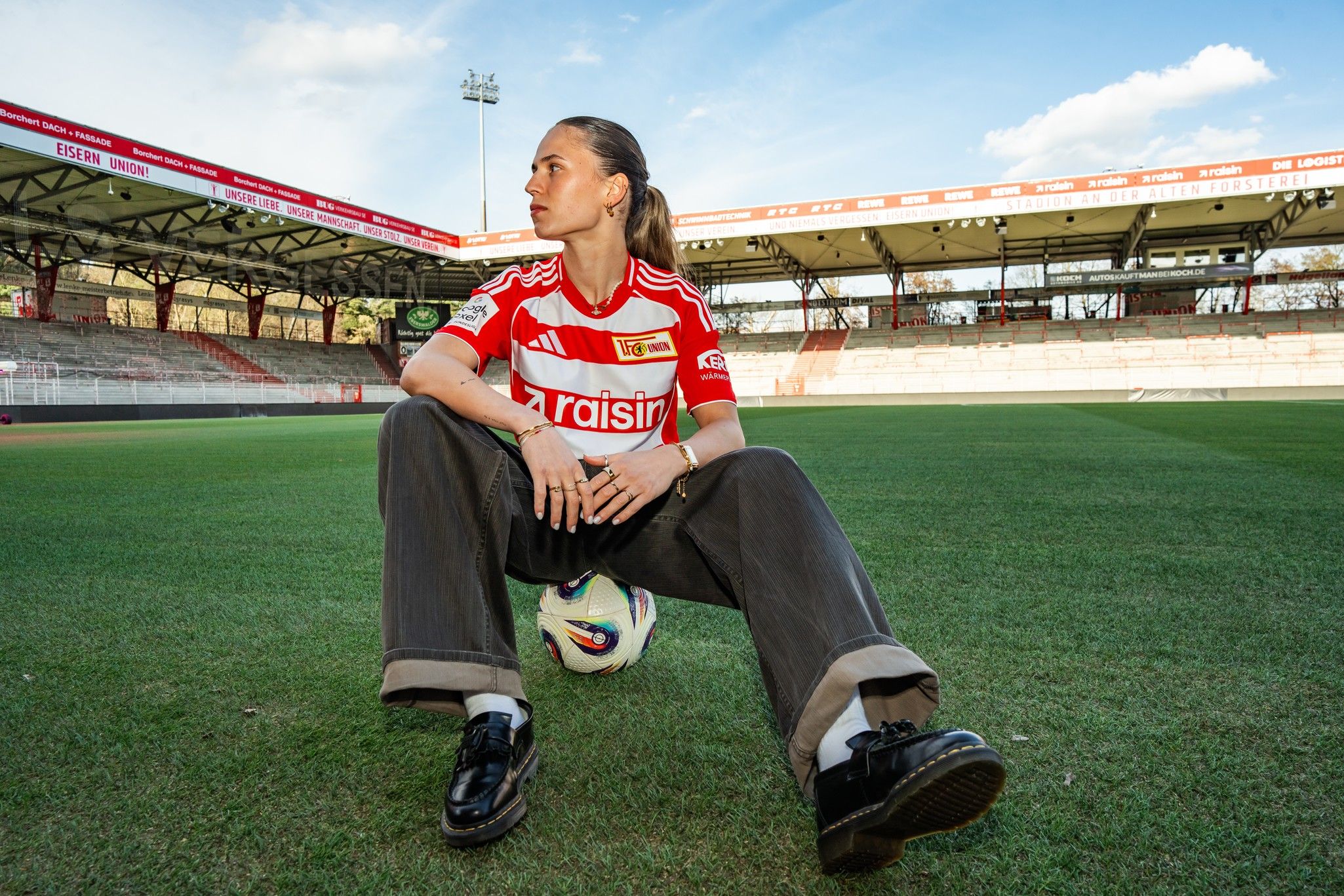Eine Person sitzt auf einem Fußball mit einem Stadion im Hintergrund, trägt ein rotes Fußballtrikot und schaut nachdenklich zur Seite.