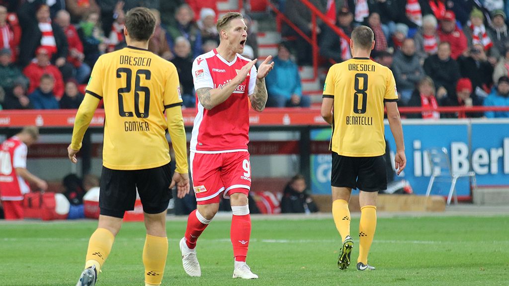 Spielszene im Fußball mit Spielern der Teams in roten und gelben Trikots auf dem Spielfeld. Zuschauer im Hintergrund.