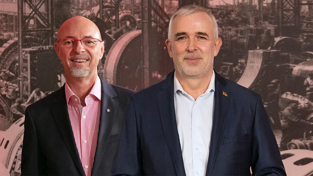 Two men in suits pose kindly in front of an industrial background with machines.