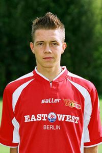 Ein junger Fußballspieler in einem roten Trikot mit dem Aufdruck „EAST WEST BERLIN“ und einem neutralen Gesichtsausdruck.