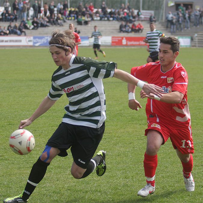 Zwei Fußballspieler im Wettkampf um den Ball auf einem Spielfeld während eines Spiels. Zuschauer sind im Hintergrund zu sehen.