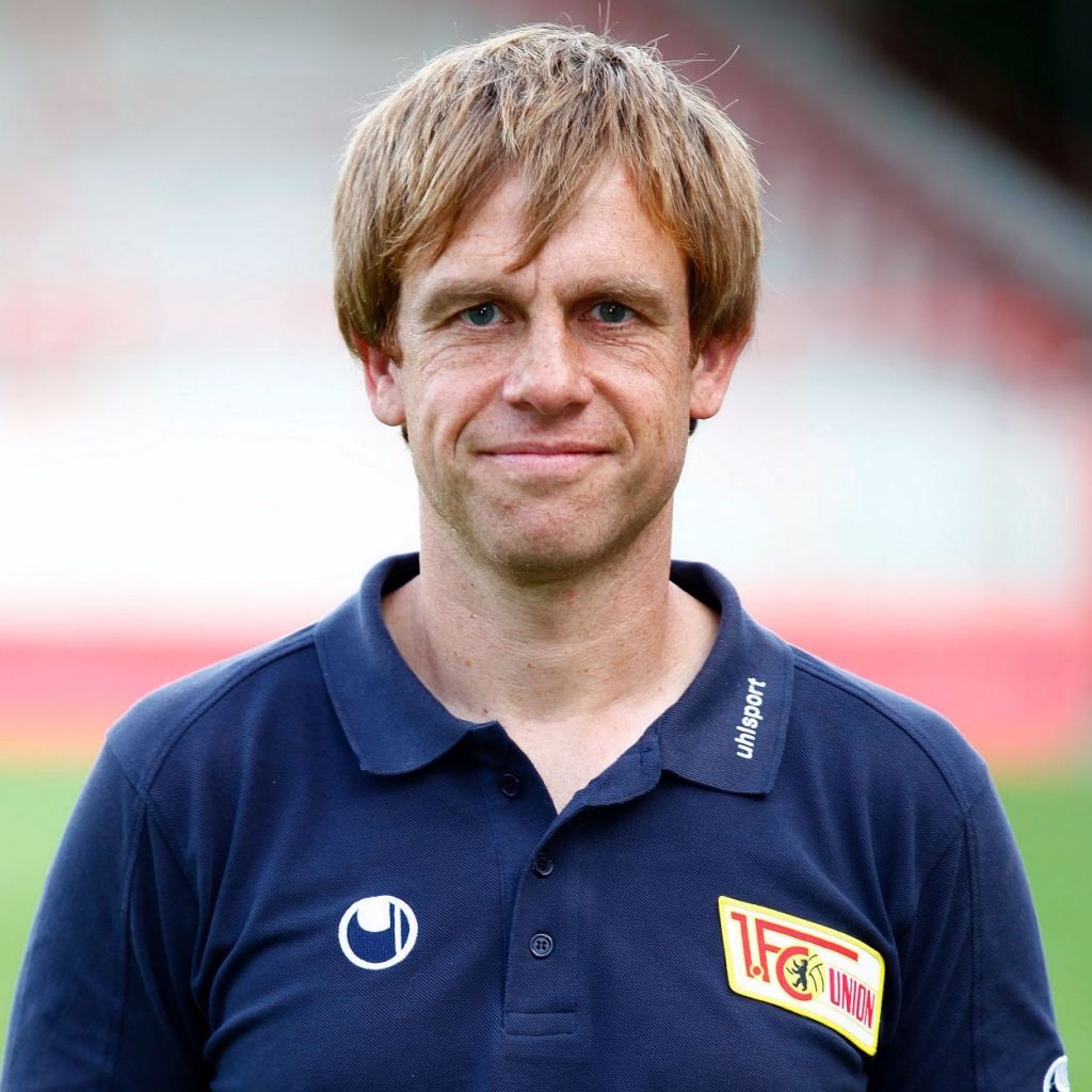 Ein lächelnder Mann in einem dunkelblauen Polo-Shirt mit dem Logo eines Fußballvereins im Hintergrund.