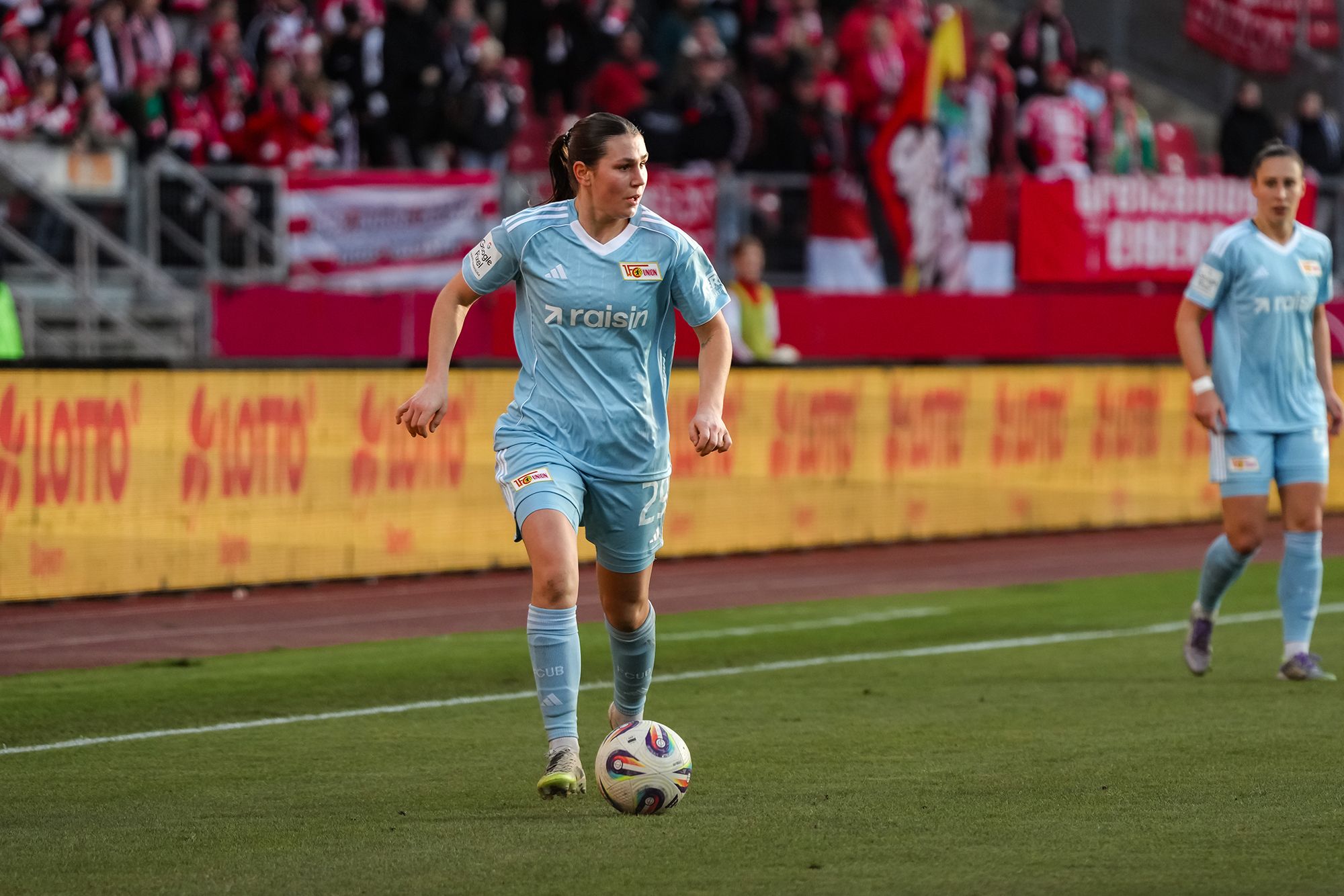 Eine Fußballspielerin in hellblauem Trikot dribbelt den Ball auf einem Stadionfeld. Zuschauer im Hintergrund.