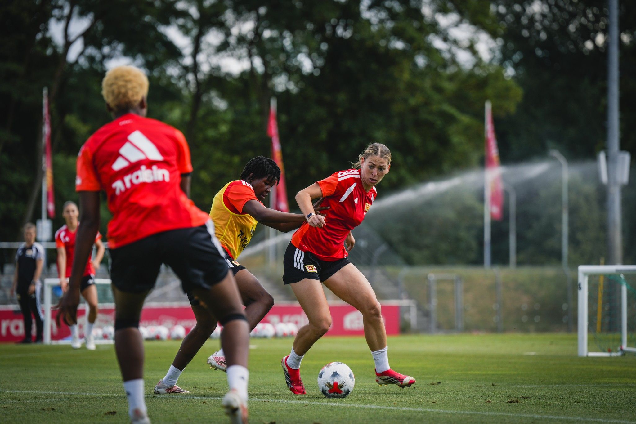 Zwei Fußballspielerinnen im Training, eine mit rotem Trikot dribbelt, während die andere sie verteidigt. Sprinkleranlagen im Hintergrund.