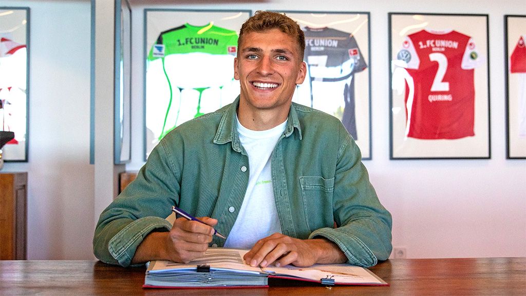 Ein lächelnder junger Mann sitzt an einem Tisch und hält einen Stift über ein offenes Buch, hinter ihm hängen Fußballtrikots.