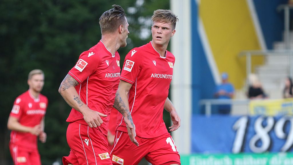 Zwei Fußballspieler in roten Trikots stehen auf dem Platz und schauen sich an, während Zuschauer im Hintergrund zu sehen sind.