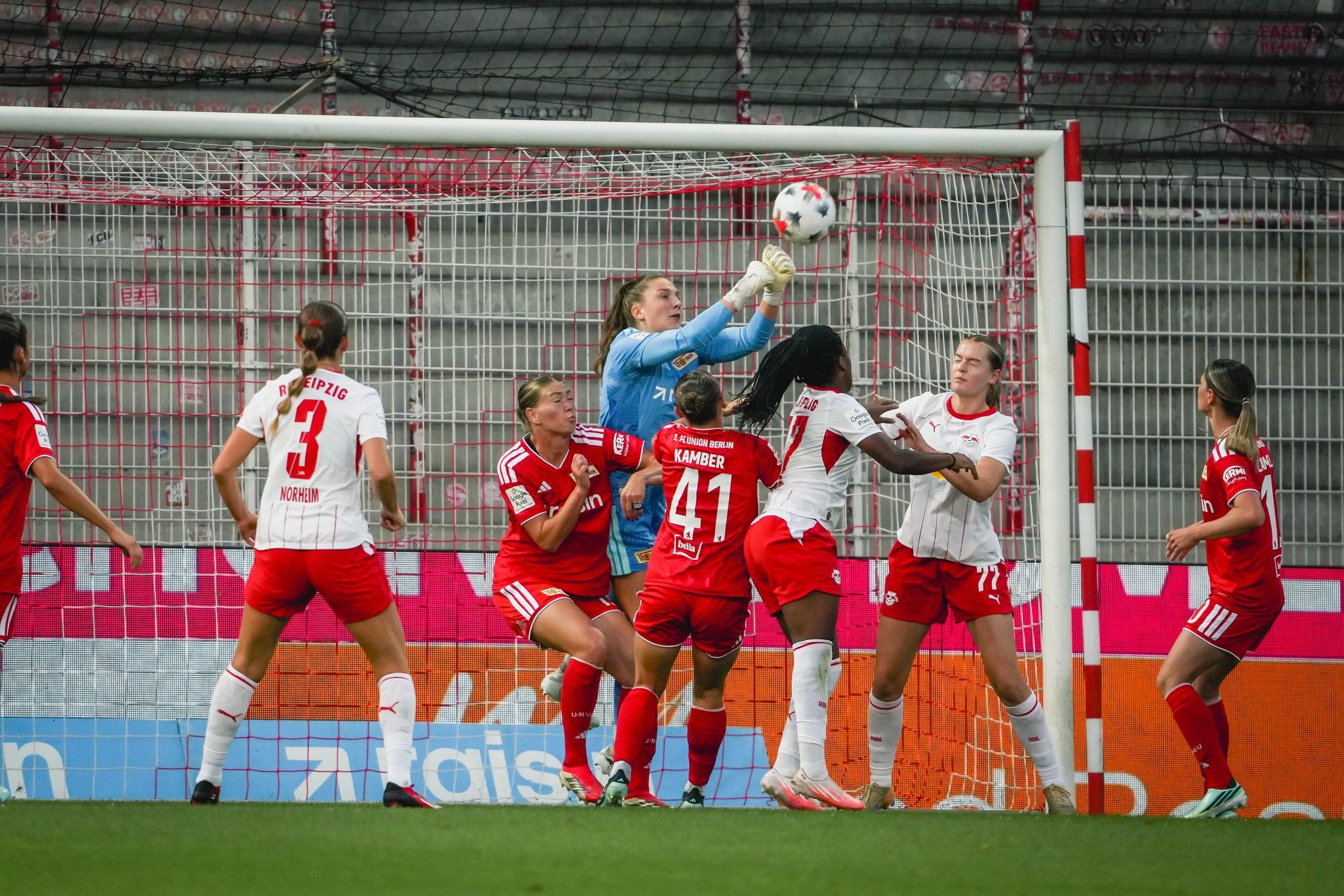 Eine Torhüterin fängt den Ball während eines Fußballspiels, umgeben von Spielerinnen des angreifenden und verteidigenden Teams.