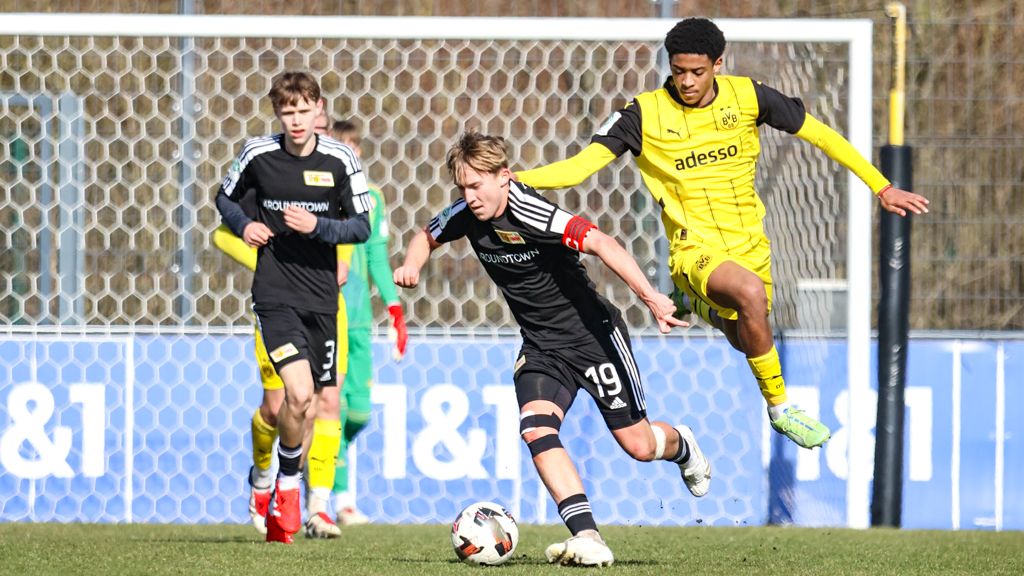 Zwei Fußballspieler verfolgen den Ball auf dem Feld, ein Spieler in Schwarz mit der Nummer 19 und ein Spieler in Gelb.