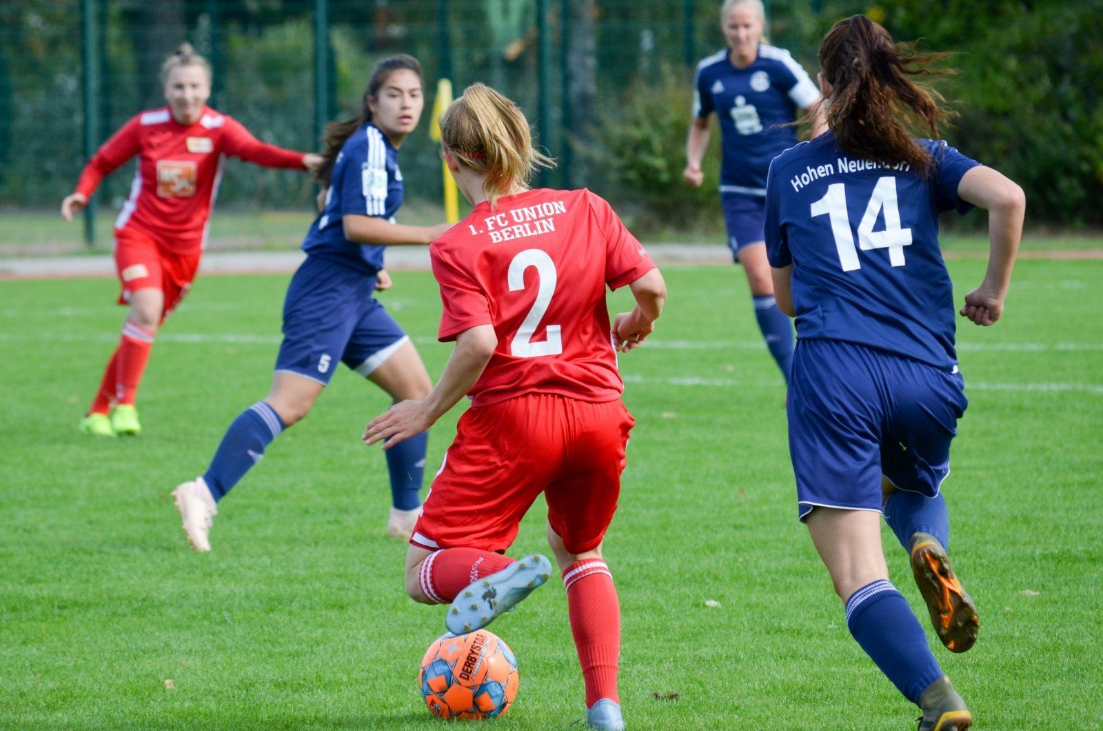 Zwei Frauenfußballmannschaften spielen auf einem grünen Platz, eine Spielerin in rotem Trikot dribbelt den Ball.
