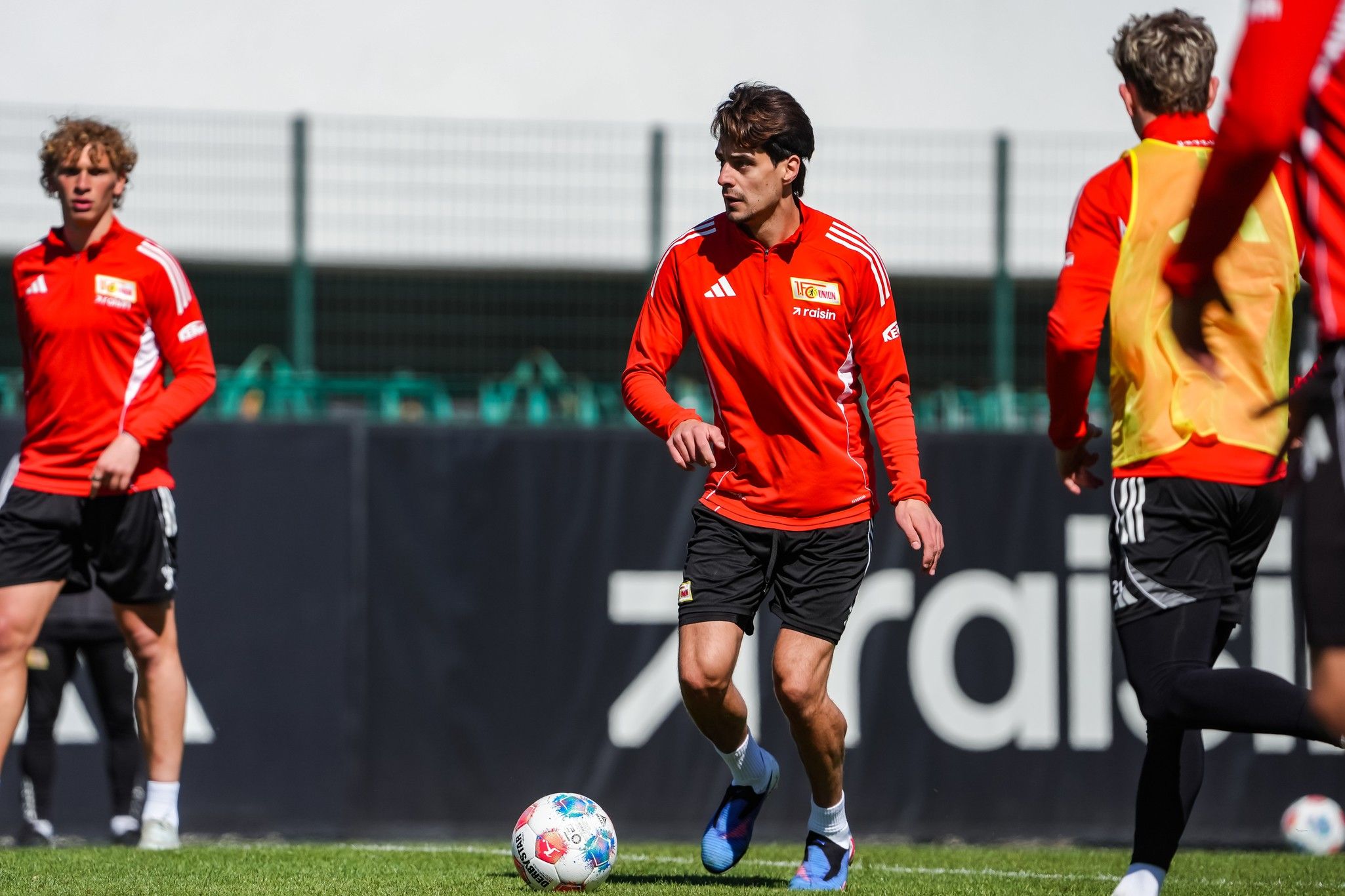Ein Fußballspieler in rotem Trainingsanzug dribbelt den Ball, während andere Spieler im Hintergrund trainieren.
