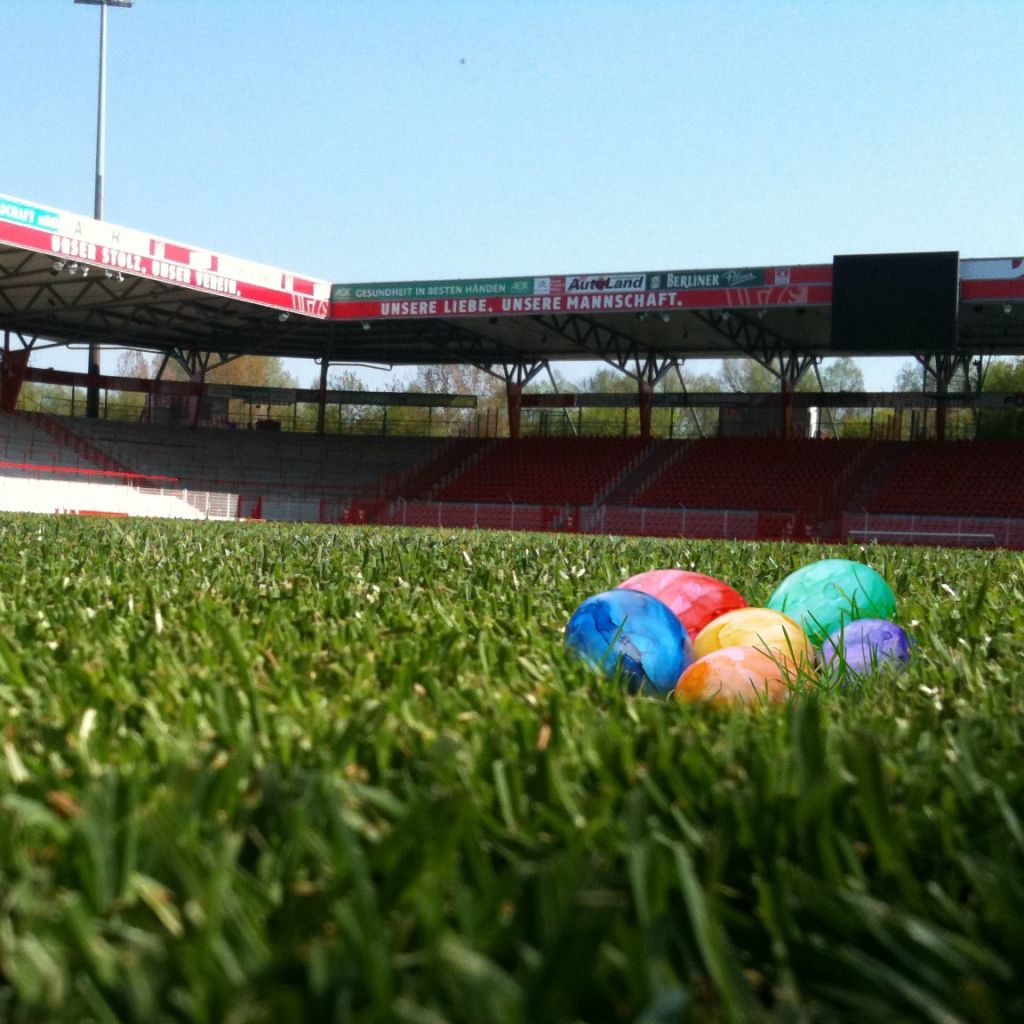 Bunte Plastikeier liegen auf einem grünen Rasen im Stadion, mit leeren Tribünen im Hintergrund und blauem Himmel.