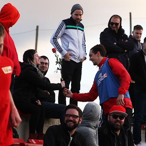 Ein Mann in Sportkleidung überreicht einer Frau mit einer Rose eine Blume, während Zuschauer im Hintergrund zuschauen.