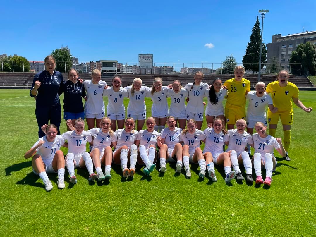 Eine Gruppe von 20 Frauen in weißen Fußballtrikots posiert freudig auf einem Rasenplatz, umgeben von Sonne und Stadthintergrund.