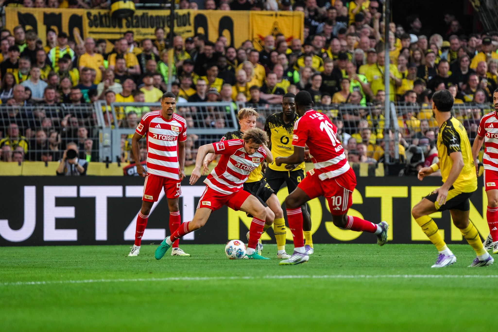 Zwei Fußballspieler kämpfen um den Ball auf dem Spielfeld, während Zuschauer im Hintergrund jubeln.