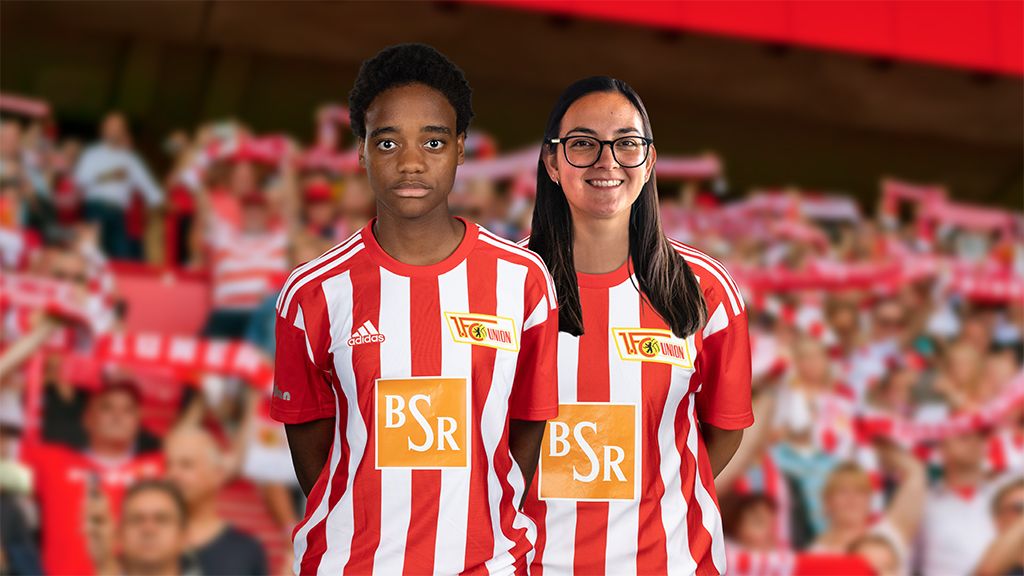 Zwei Personen in rot-weiß gestreiften Trikots stehen vor verschwommenen Fans im Hintergrund.