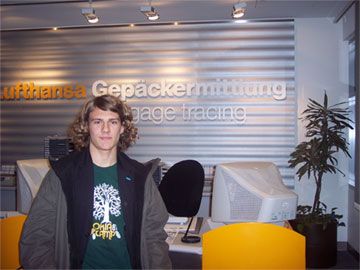 Junger Mann mit lockigem Haar steht vor einem Empfangstresen mit dem Schriftzug "Lufthansa Gepäckermittlung".