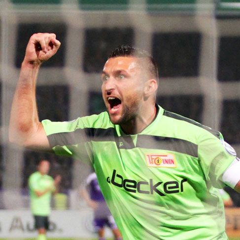 Ein jubelnder Fußballspieler in grüner Torwartkleidung feiert begeistert einen Erfolg auf dem Spielfeld.