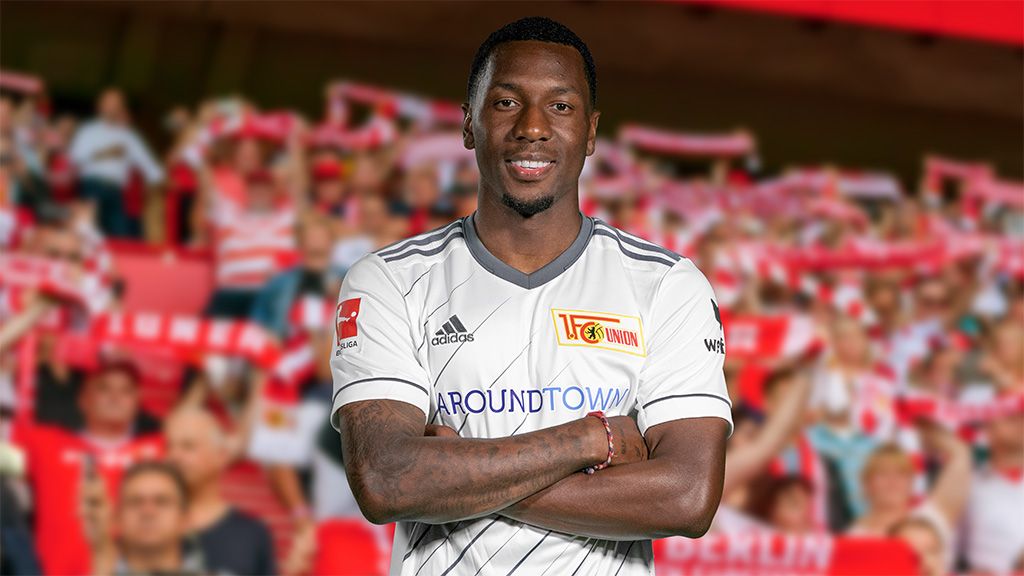 Ein Fußballspieler in einem weißen Trikot mit Vereinslogo steht lächelnd vor jubelnden Fans im Stadion.