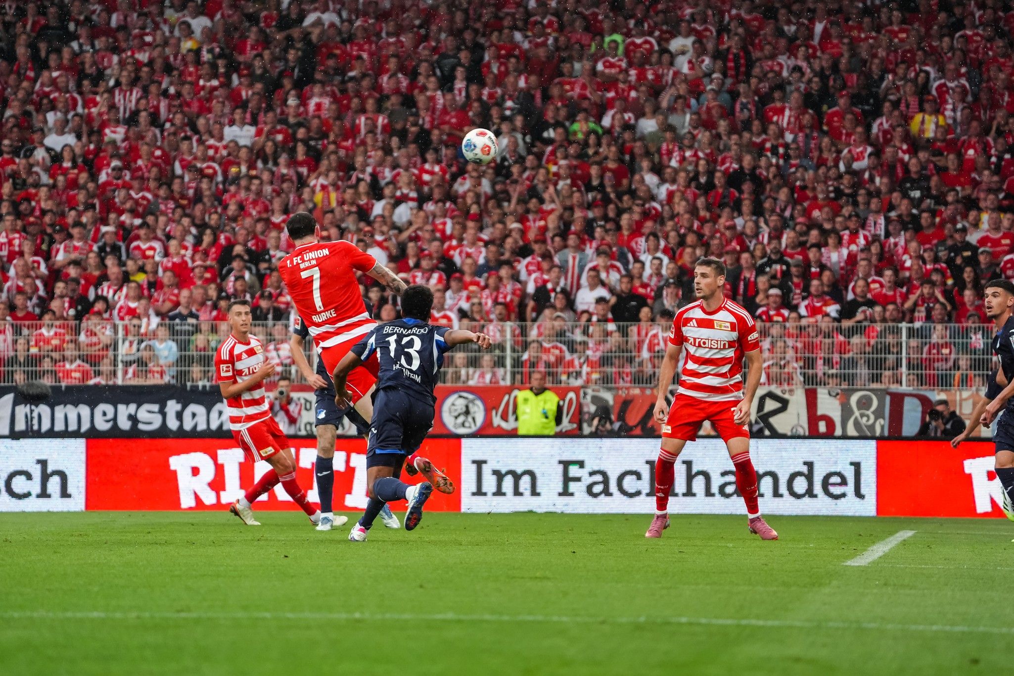 Ein Fußballspieler springt hoch, um einen Ball zu köpfen, während Zuschauer in roten Trikots jubeln.