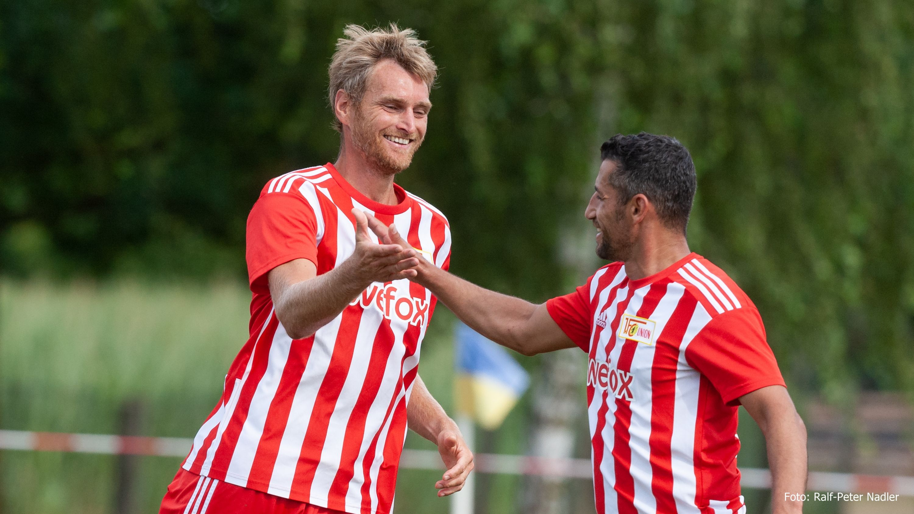 Zwei Fußballspieler in rot-weißen Trikots jubeln sich gegenseitig zu, umgeben von einer grünen Landschaft.