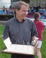 Ein Mann steht auf einem Sportfeld und hält ein Tablett mit einer mit Stäben versehenen Speise in der Hand, während er lächelt.