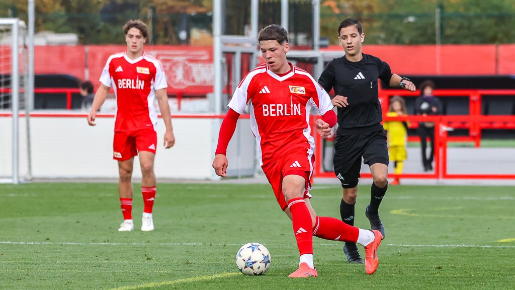 Ein Fußballspieler in rotem Trikot mit der Aufschrift "BERLIN" dribbelt den Ball auf dem Spielfeld, zwei Mitspieler sind im Hintergrund.
