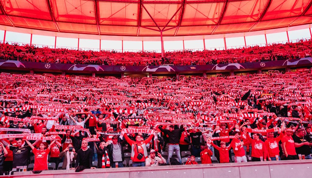 Ein Stadion voller jubelnder Fans in roten Trikots und Schals, die ihre Mannschaft unterstützen.