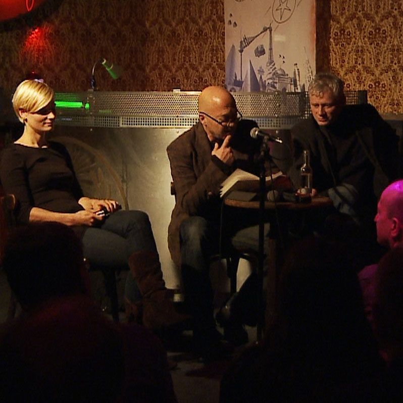 Drei Personen sitzen auf einer Bühne, während ein Mann in der Mitte vor einem Mikrofon liest. Hintergrund mit Dekoration und schwachem Licht.