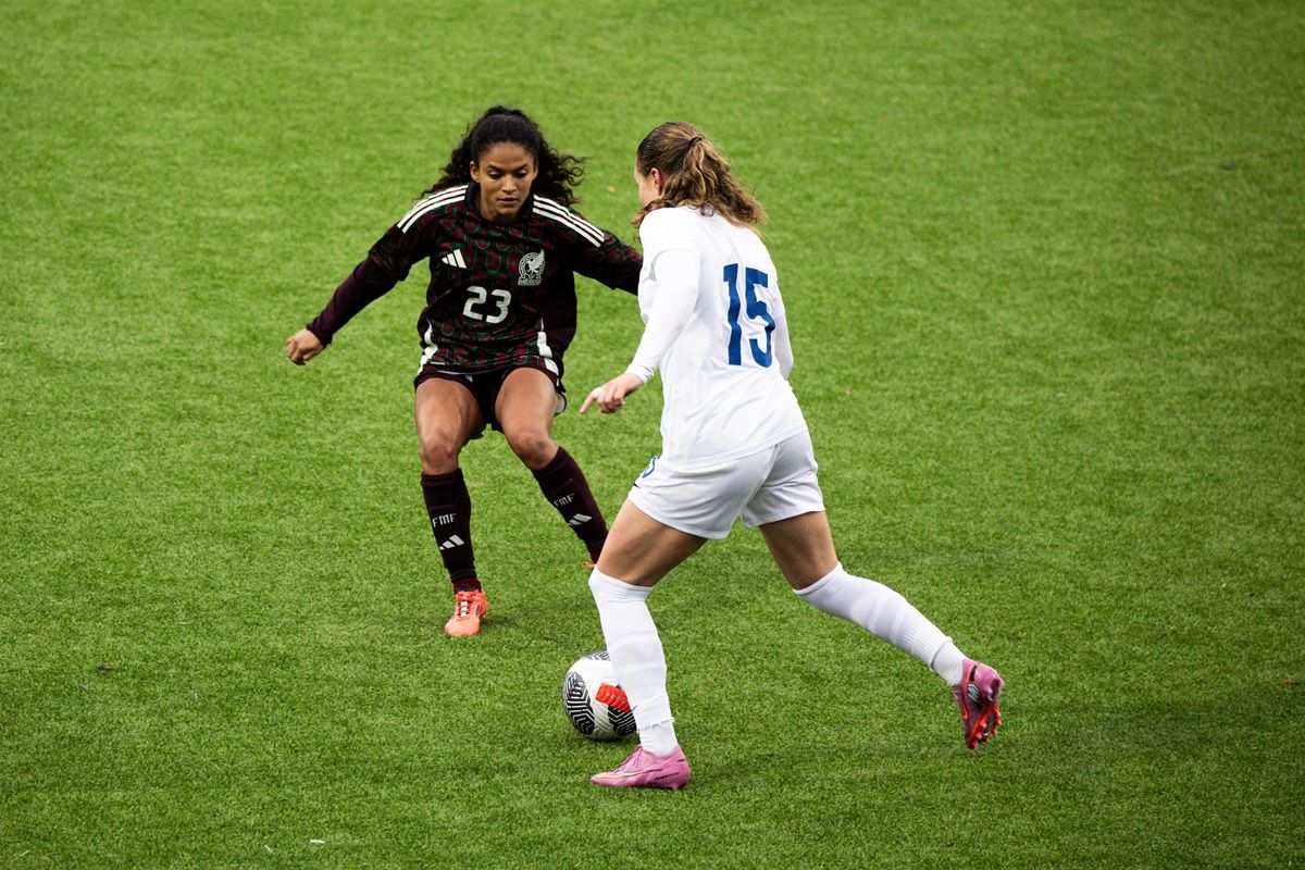 Eine mexikanische Fußballspielerin mit der Nummer 23 dribbelt gegen eine Spielerin mit der Nummer 15 in einem weißen Trikot auf dem Spielfeld.