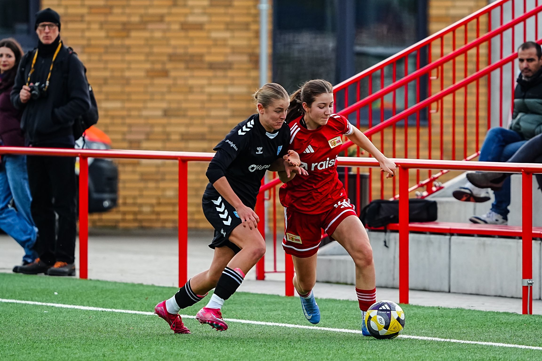Zwei Fußballspielerinnen spielen auf einem Platz. Sie kämpfen um den Ball. Im Hintergrund stehen Zuschauer.
