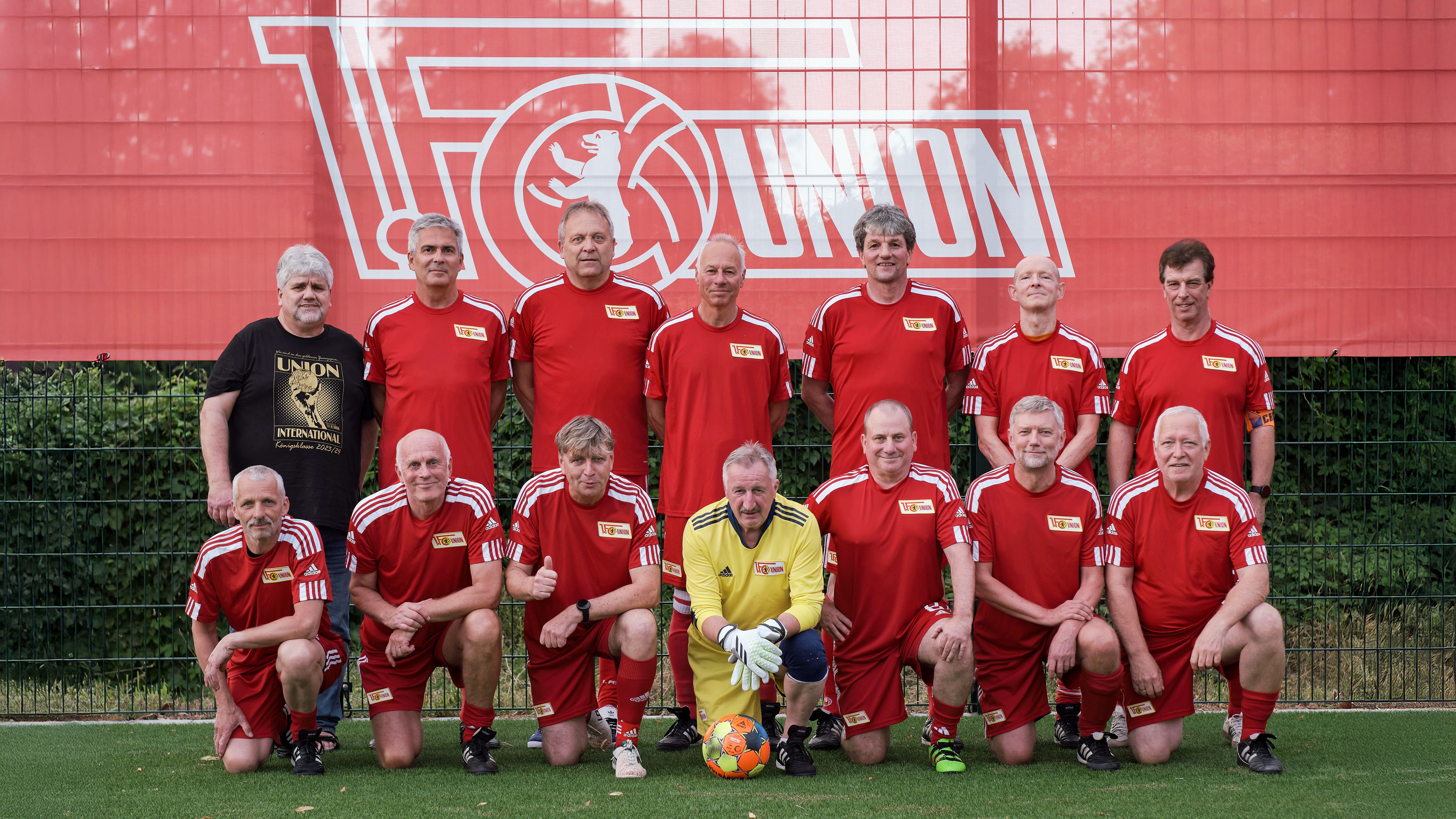 Eine Gruppe älterer Männer in roten Fußballtrikots steht auf einem Sportplatz vor einem roten Banner mit dem Schriftzug "FC Union".