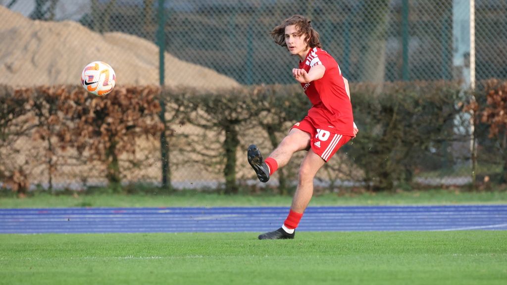 Ein junger Fußballspieler in rotem Trikot schießt den Ball auf einem Rasenplatz. Im Hintergrund sind Bäume und eine Sandschicht sichtbar.