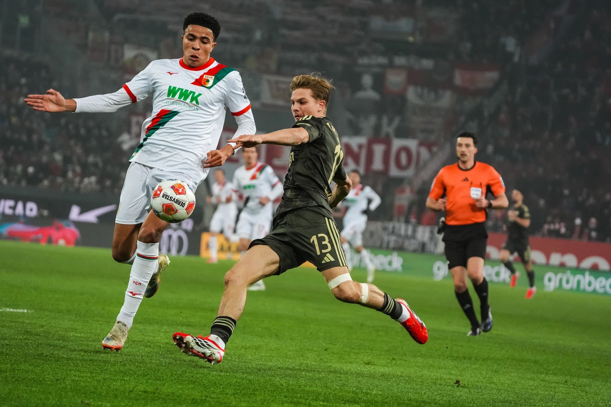 Zwei Fußballspieler in Aktion, während einer den Ball spielt und der andere versucht, ihn zu verteidigen. Im Hintergrund Zuschauer und ein Schiedsrichter.