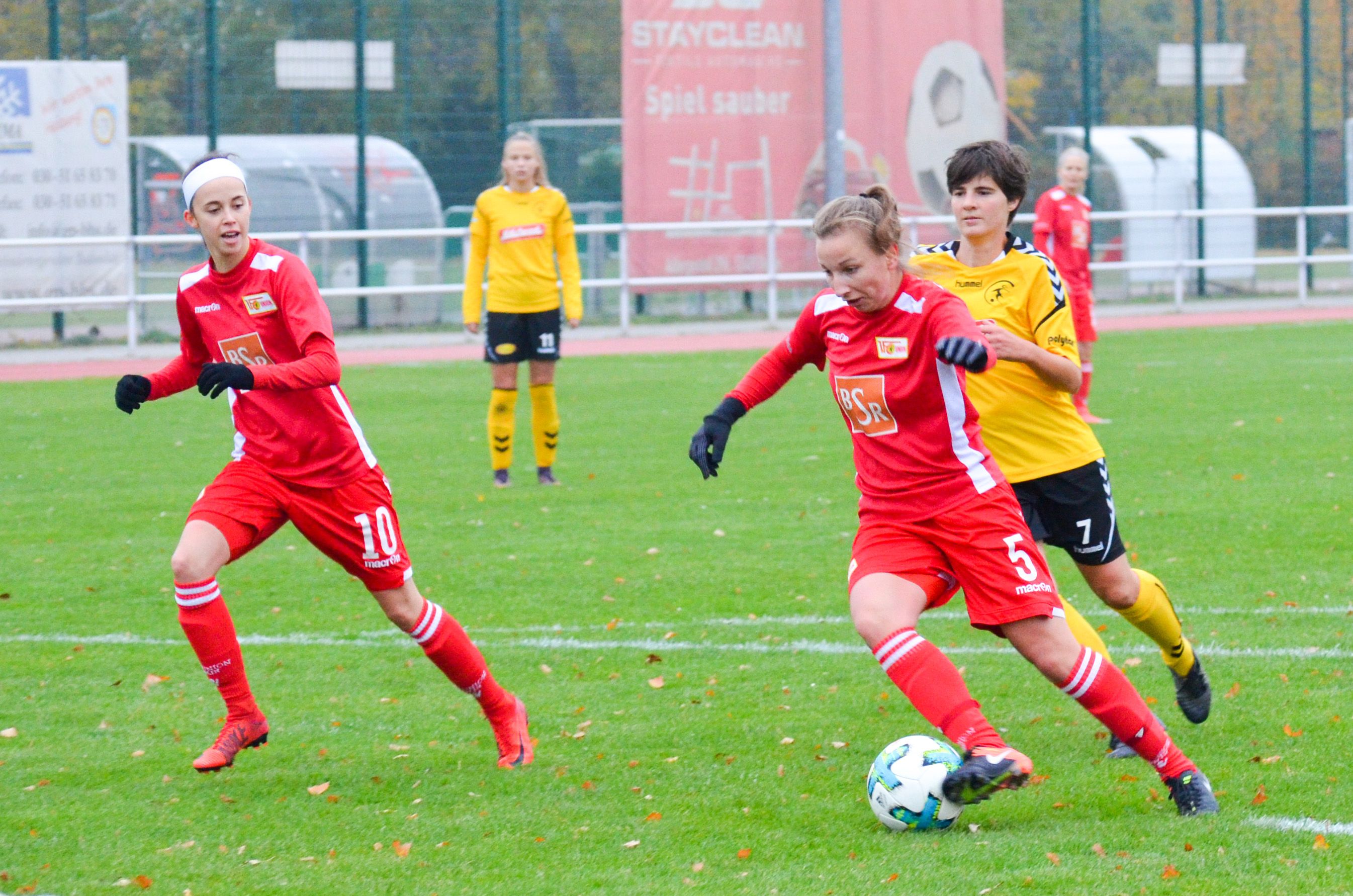 Eine Fußballspielszene mit zwei Spielerinnen in roten Trikots und einer in gelbem Trikot, die um den Ball kämpfen.