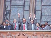 Eine Gruppe von Männern in Anzügen jubelt und hebt die Arme auf einem Balkon vor einem historischen Gebäude.