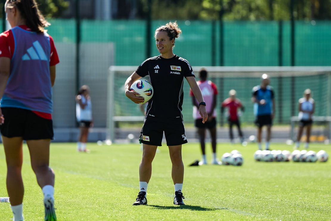 Eine weibliche Trainerin steht auf einem Fußballplatz und hält einen Ball in der Hand, umgeben von Spielern im Training.