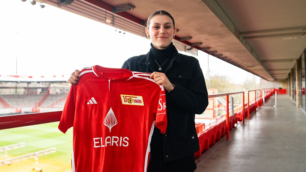 A person is holding a red soccer jersey with a club's logo in a stadium.