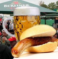 Ein Bierkrug und ein Brötchen mit Bratwurst auf einer Veranstaltung im Freien, mit Menschen im Hintergrund.