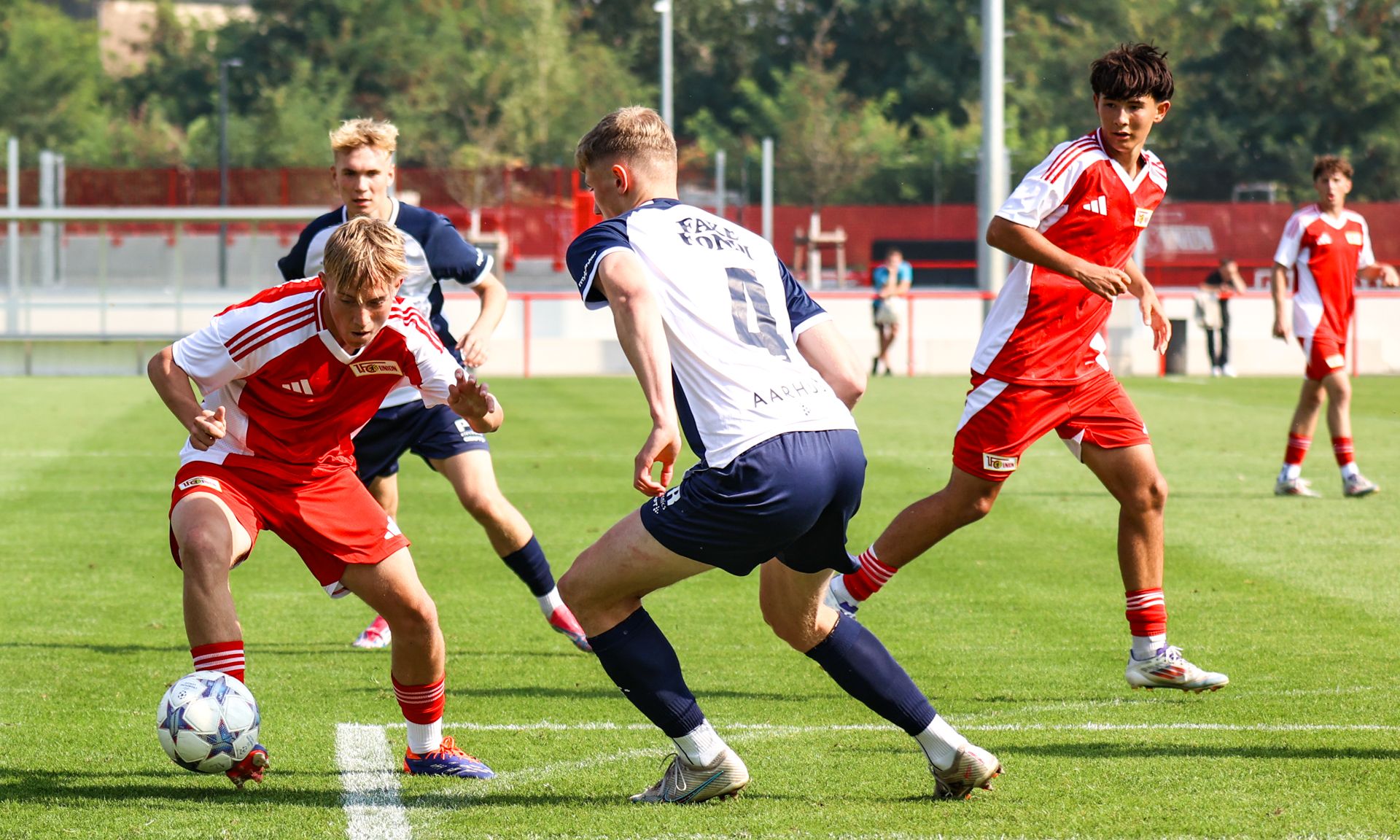 Junge Fußballspieler in roten und blauen Trikots kämpfen um den Ball auf einem grünen Rasenplatz.