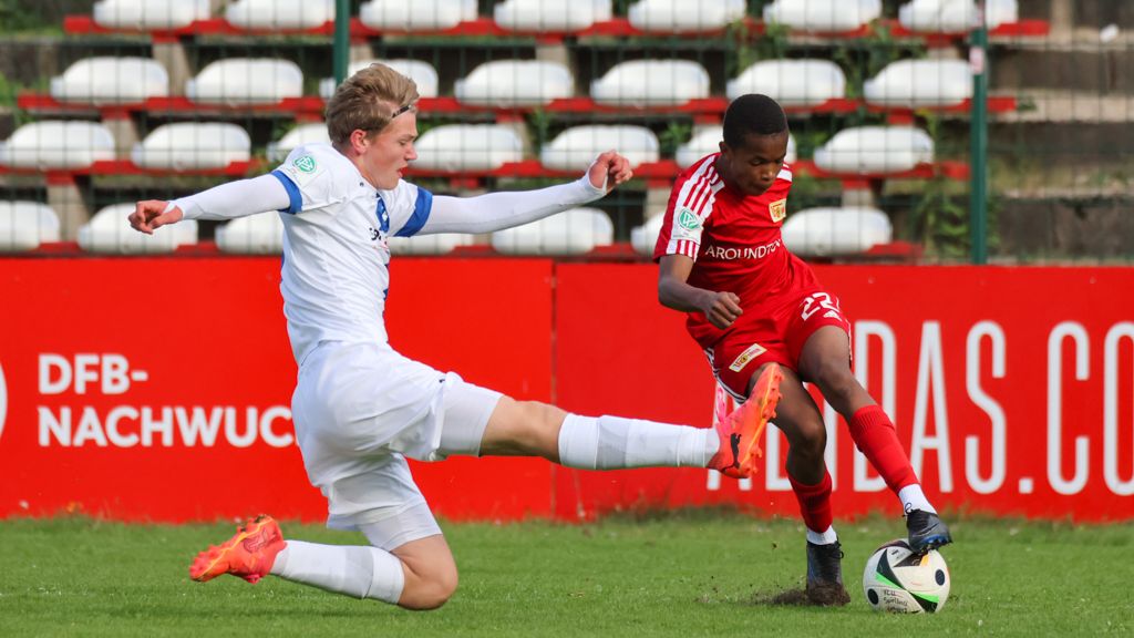 Ein junger Fußballspieler in roter Ausrüstung dribbelt, während ein Gegner in weißem Trikot versucht, ihn zu stoppen.