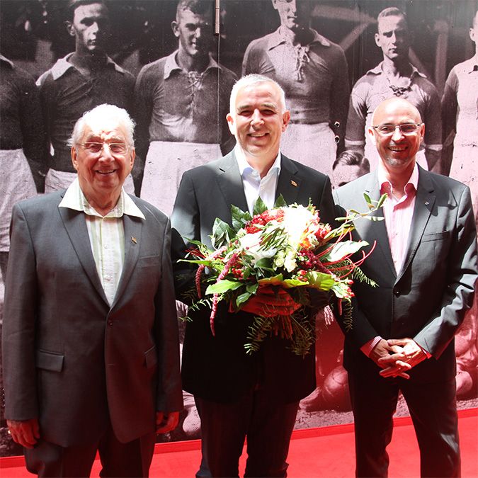 Drei Männer stehen lächelnd auf einem roten Teppich, umgeben von Blumen und einer historischen Fotografie im Hintergrund.